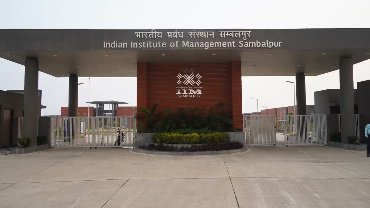 The main entrance gate of IIM Sambalpur, bilingual signage in Hindi and English on a wide horizontal lintel above a central pavilion with the IIM Sambalpur logo on red brick, flanked by open gates and the campus driveway.
