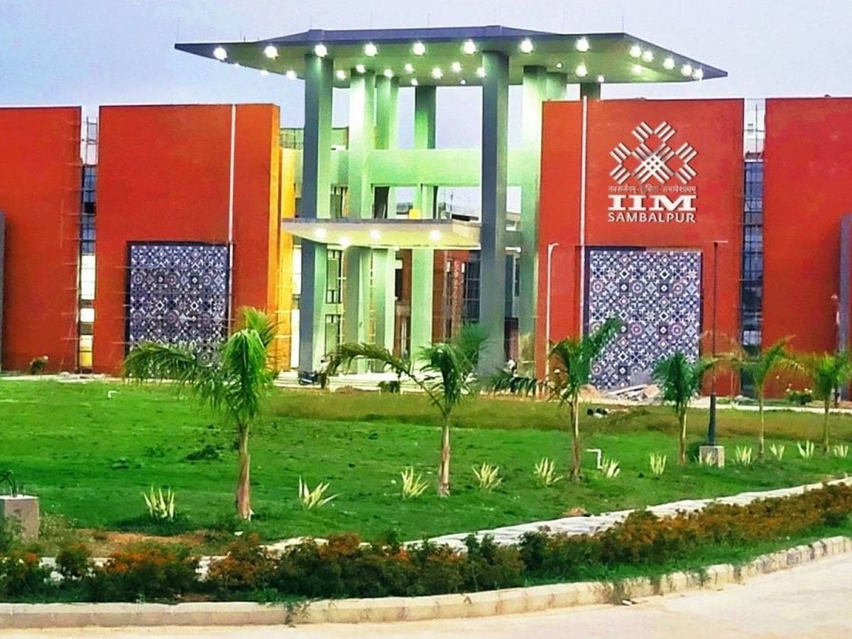 Respresentative Image. The main entrance gateway of IIM Sambalpur, terracotta-red flanking blocks with Sambalpuri ikat-pattern decorative panels, central green pavilion crowned with overhead lighting, and the IIM Sambalpur logo on the right wall.