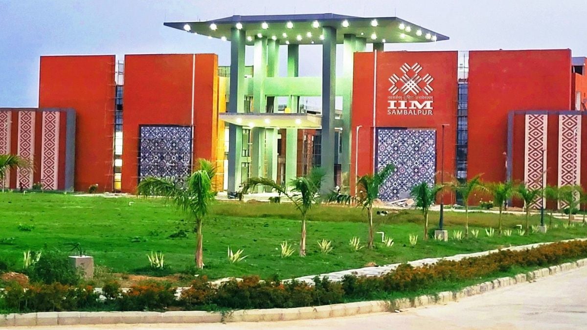 The main entrance gateway of IIM Sambalpur, terracotta-red flanking blocks with Sambalpuri ikat-pattern decorative panels, central green pavilion crowned with overhead lighting, and the IIM Sambalpur logo on the right wall.
