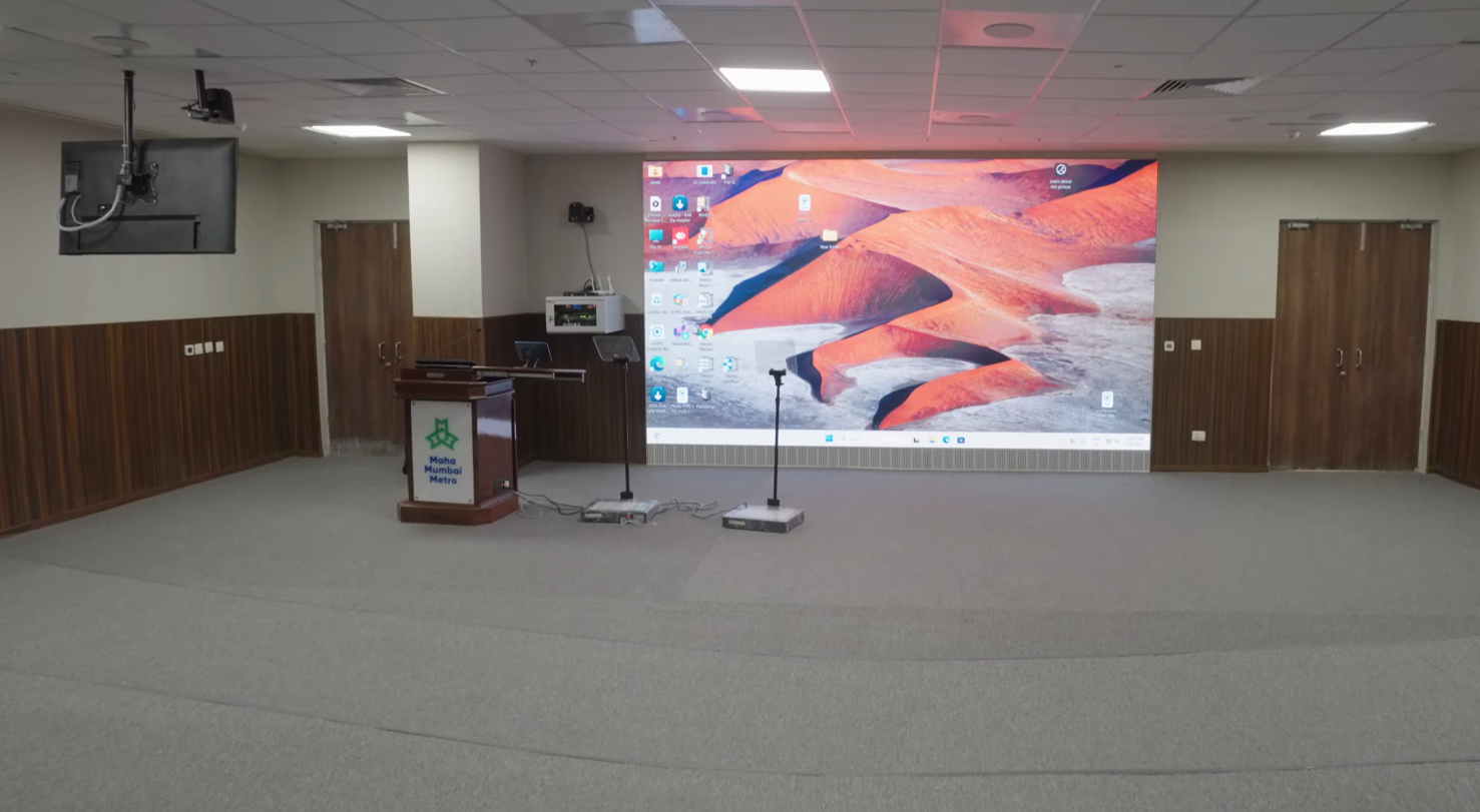 One of the two hybrid auditoriums at MMTI, photographed from the rear of the room with a wall-spanning LED video display and presentation rostrum at the front, acoustic wood and fabric panelling on the surround walls, ceiling-mounted projector and beamforming devices overhead.