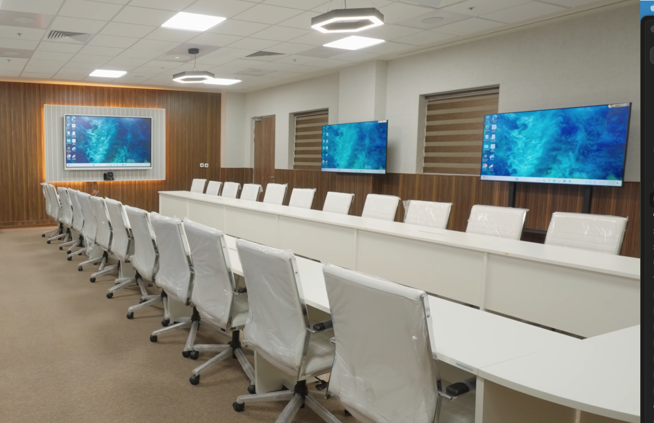 A long boardroom-style hybrid conference room at MMTI with a single principal interactive flat panel at the head of the table, two confidence displays mounted on the side wall, white meeting table with chairs around it, wood-panelled dado, decorative hexagonal pendant lighting, and a busy ceiling carrying recessed lighting and HVAC return slots.